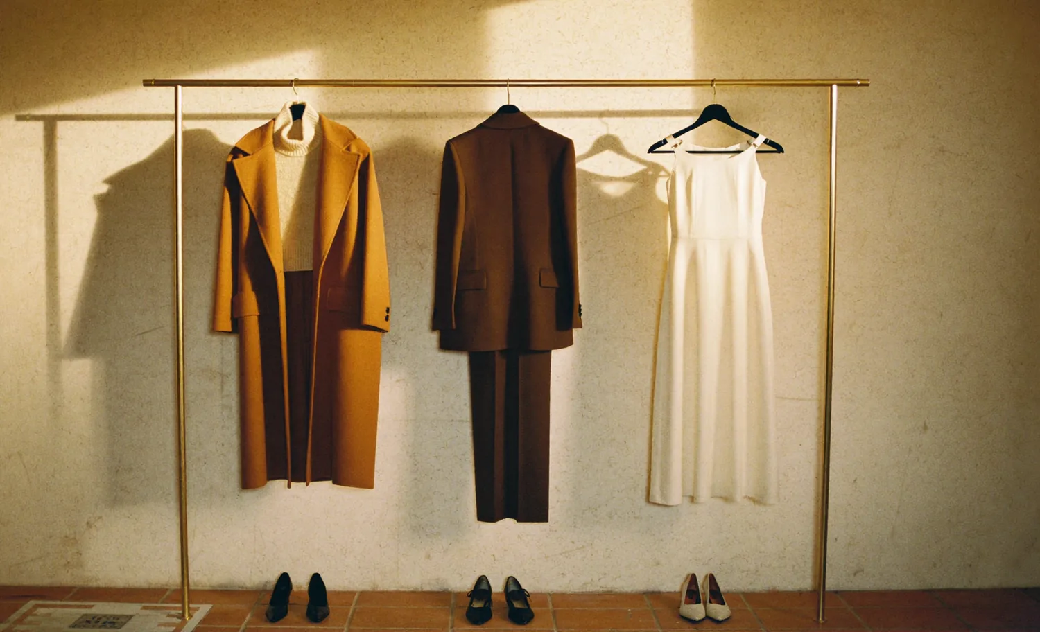 Three complete outfits on a brass rail, shoes placed beneath each, in warm afternoon light.